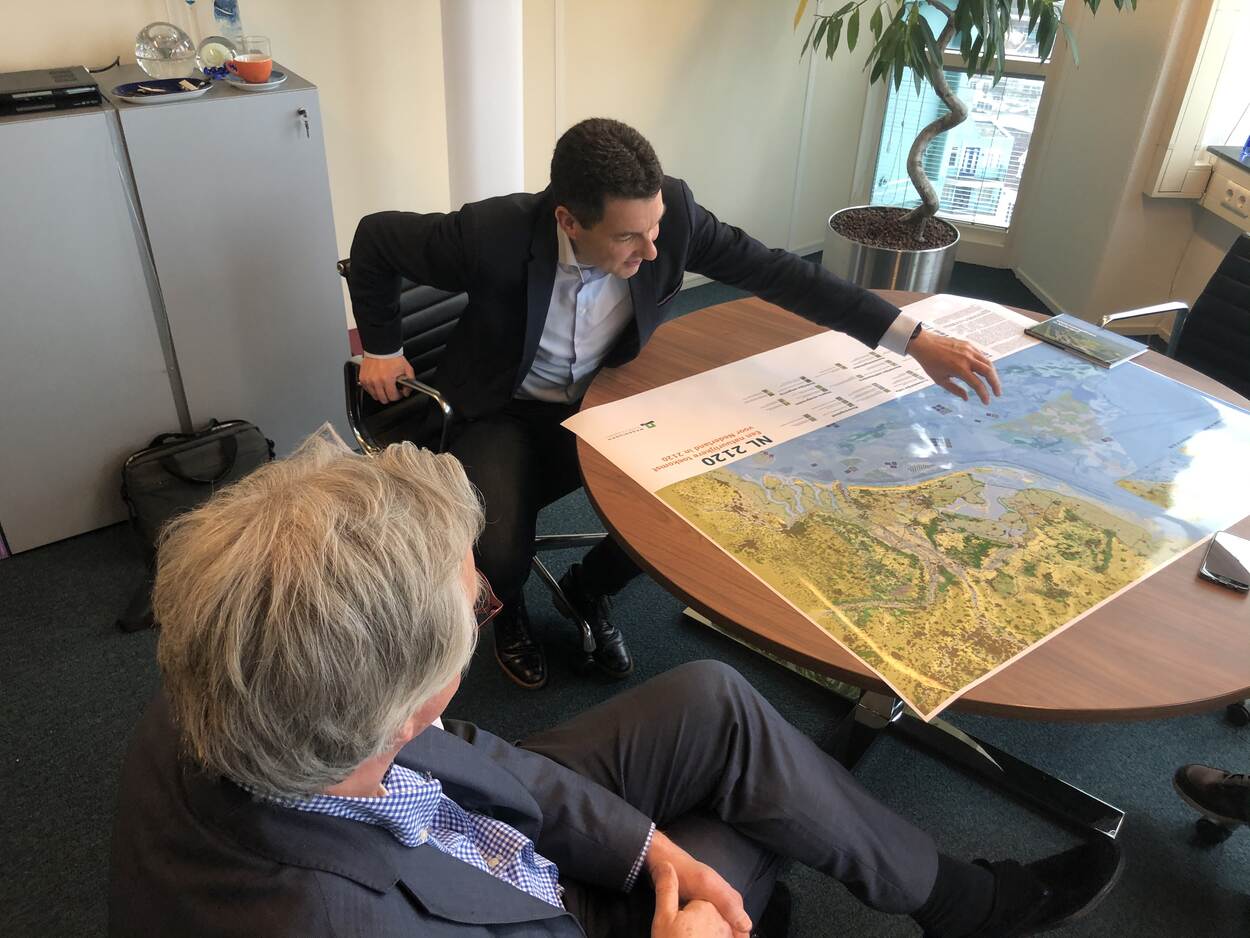 Deltacommissaris Peter Glas (links op de foto) en Tim van Hattum (rechts op de foto) zitten aan een tafel. Op de tafel ligt een grote kaart van Nederland. Van Hattum wijst iets aan op de kaart.