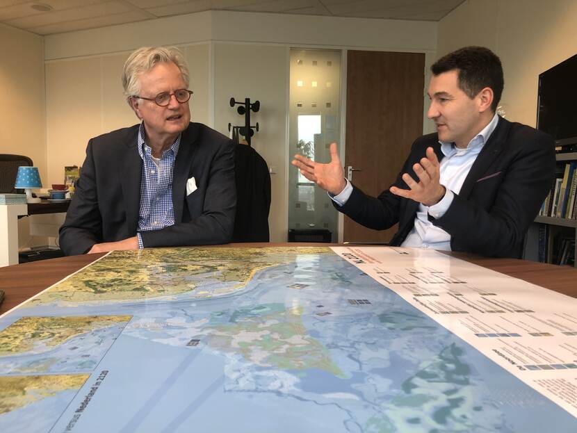 Deltacommissaris Peter Glas (links op de foto) en Tim van Hattum (rechts op de foto) zitten aan een tafel. Op de tafel ligt een grote kaart van Nederland.