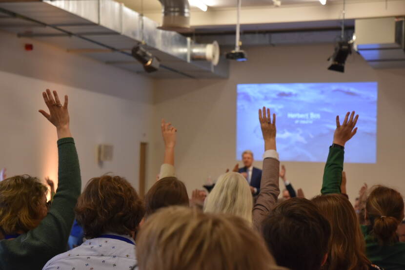 De bijeenkomst vanachter het publiek gefotografeerd, Een viertal deelnemers steekt hun hand op.