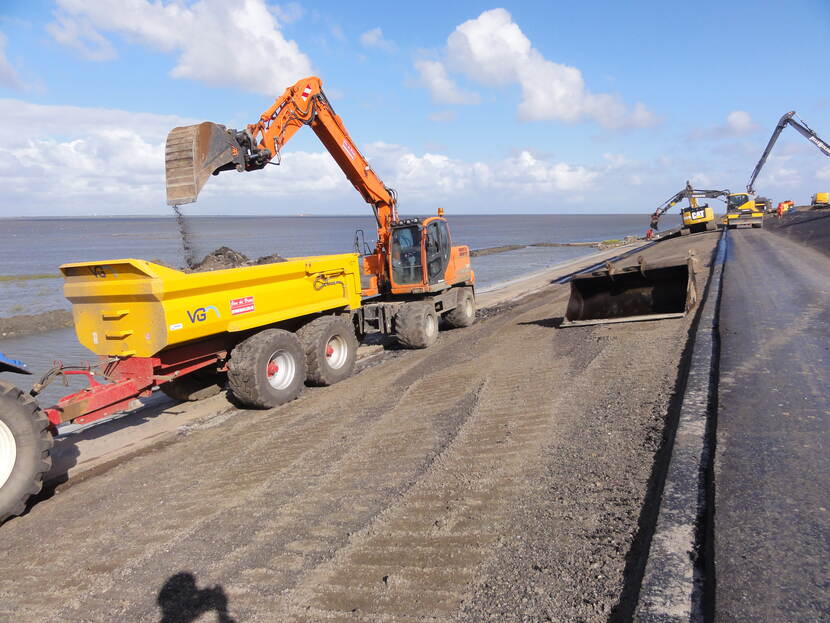 Verschillende oranje-gele graafmachines werken op een dijk. Op de dijk ligt asfalt en aan de linkerzijde van de dijk is water te zien.