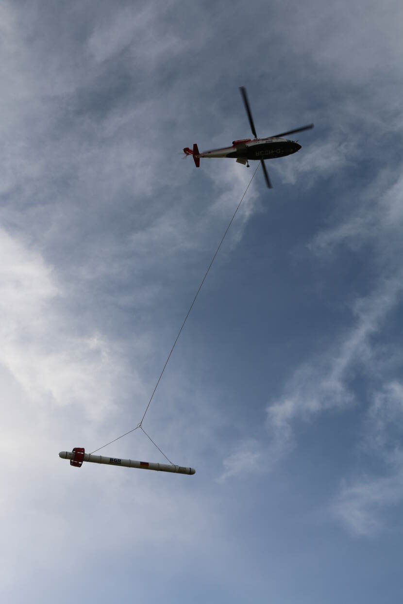Vliegende helicopter van onder gefotografeerd. Aan de helicopter hangt touw met aan het uiteinde een paal.
