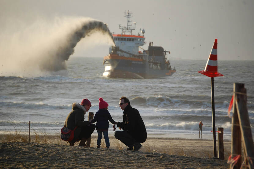 Foto van schip dat zand opspuit op de achtergrond. Op de voorgrond een gezinnetje dat naar het strand gaat.