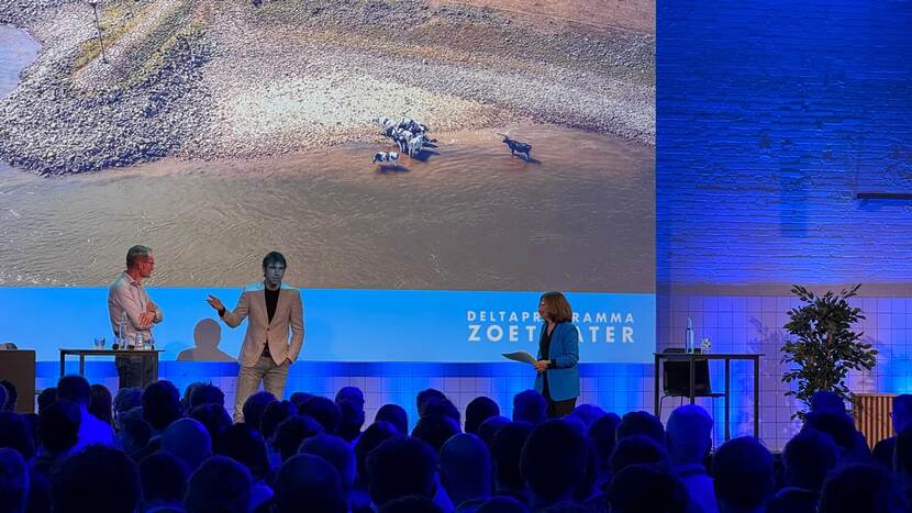 Drie mensen tijdens een presentatie, in een zaal vol mensen.