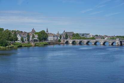 Een zonnige dag in Maastricht. We zien de maas met in de verte een oude brug met bogen. We zien een stuk van de stad vanaf de kade aan de linkerkant van de foto.
